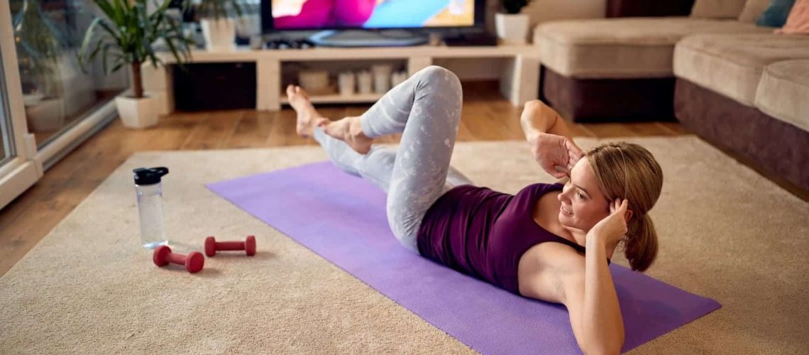 mulher-atletica-feliz-praticando-abdominais-enquanto-se-exercita-na-frente-de-uma-tv-em-casa (Grande)